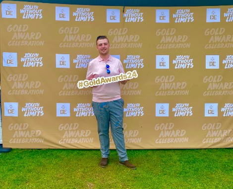 A male stood in front of a gold Duke of Edinburgh award backdrop holding a GoldAwards24 sign.